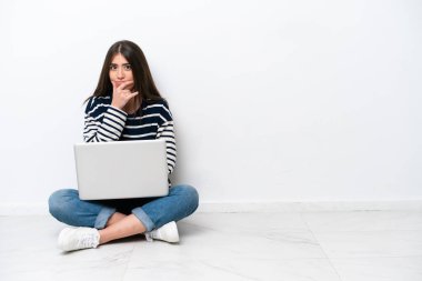 Yerde dizüstü bilgisayarlı beyaz bir kadın oturuyor. Beyaz arka planda izole edilmiş. Şaşırmış ve şoka uğramış.