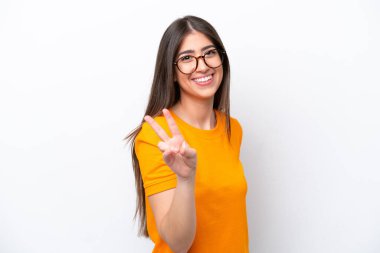 Beyaz arka planda izole edilmiş genç beyaz kadın gülümsüyor ve zafer işareti gösteriyor.