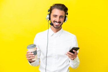 Tele pazarlamacı beyaz adam sarı arka planda izole edilmiş bir kulaklıkla çalışıyor. Elinde kahve ve cep telefonu var.