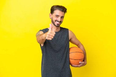 Yakışıklı genç basketbolcu sarı arka planda izole edildi. Başparmakları havada çünkü iyi bir şey oldu.