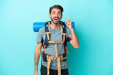 Mavi arka planda izole edilmiş büyük bir sırt çantasıyla genç dağcı bir parmağını kaldırarak çözümü anlamaya çalışıyor.