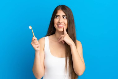 Mavi arka planda dişlerini fırçalayan genç beyaz kadın yukarı bakarken bir fikir düşünüyor.