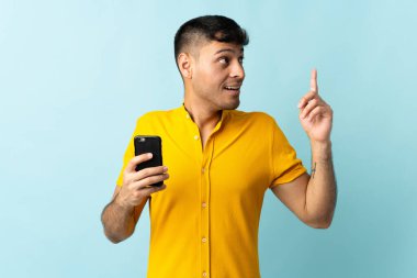 Cep telefonu kullanan genç Kolombiyalı adam mavi arkaplandan izole edilmiş. Çözümü bulmak için parmağını kaldırıyor.