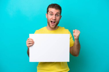 Kafkasyalı genç adam mavi arka planda izole edilmiş boş bir levha tutuyor ve zaferi kutluyor.