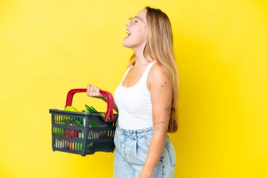 Sarı arka planda izole edilmiş bir alışveriş sepeti taşıyan beyaz bir kadın yan yana gülerek gülüyor.