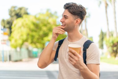 Genç yakışıklı bir adam açık havada kahvesini yudumlarken bir fikir üretiyor ve etrafına bakıyor.
