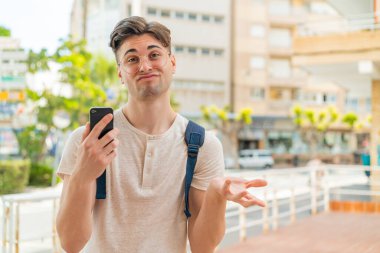 Genç yakışıklı adam dışarıda cep telefonu kullanıyor. Omuzlarını kaldırırken şüpheli hareketler yapıyor.