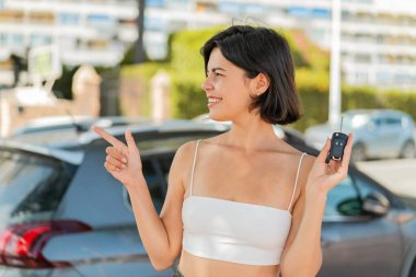 Genç, güzel bir Bulgar kadın dışarıda araba anahtarı tutuyor ve bir ürün sunmak için kenarı işaret ediyor