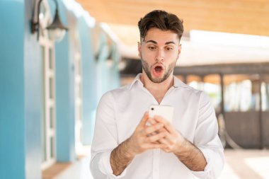 Genç yakışıklı adam kameraya bakarken şaşırmış bir ifadeyle cep telefonunu kullanıyor.