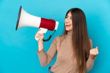 Mavi arka planda izole edilmiş genç beyaz bir kadın megafondan bir şey duyurmak için bağırıyor.