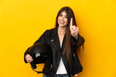 Sarı arka planda motosiklet kaskı tutan genç beyaz kadın parmağını kaldırıyor ve gösteriyor.