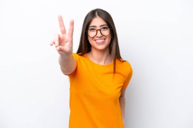 Beyaz arka planda izole edilmiş genç beyaz kadın gülümsüyor ve zafer işareti gösteriyor.