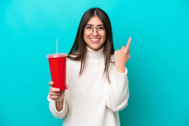 Mavi arka planda izole edilmiş gazoz içen genç beyaz kadın en iyinin işareti olarak parmağını kaldırıyor.