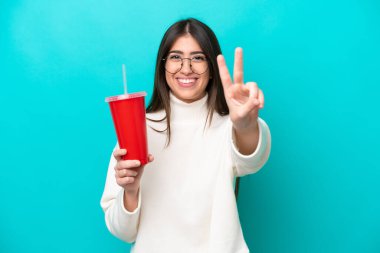 Mavi arka planda gazoz içen genç beyaz kadın gülümsüyor ve zafer işareti gösteriyor.