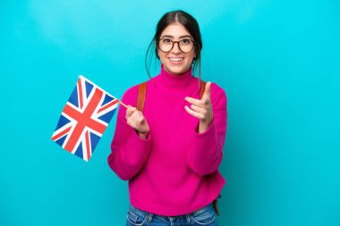 Mavi arka planda İngiliz bayrağı taşıyan genç beyaz kadın şaşırmış ve ön tarafı işaret ediyor.