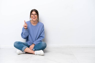 Beyaz arka planda izole bir şekilde yerde oturan beyaz tenli genç kadın çözümün farkına varmak için parmağını kaldırıyor.