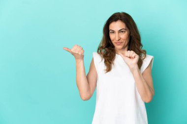 Mavi arka planda izole edilmiş orta yaşlı beyaz kadın bir ürünü sunmak için kenarı işaret ediyor.