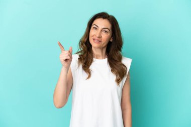 Mavi arka planda izole edilmiş orta yaşlı beyaz kadın en iyinin işareti olarak parmağını kaldırıyor.