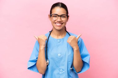 Genç Kolombiyalı hemşire pembe arka planda izole edilmiş. Baş parmakları havada ve gülümsüyor.