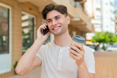 Genç yakışıklı adam cep telefonu kullanıyor ve elinde kahve tutuyor.