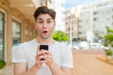 Genç yakışıklı adam kameraya bakarken şaşırmış bir ifadeyle cep telefonunu kullanıyor.