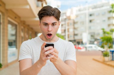 Genç yakışıklı adam kameraya bakarken şaşırmış bir ifadeyle cep telefonunu kullanıyor.