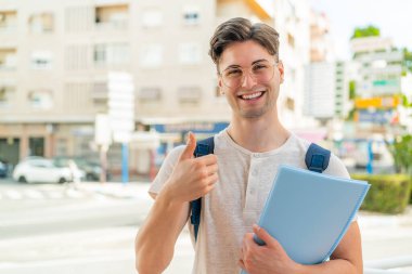 Açık havada genç ve yakışıklı bir öğrenci. Başparmakları havada. Çünkü iyi bir şey oldu.