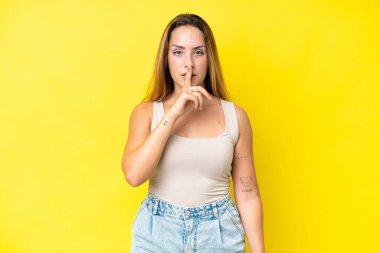 Sarı arka planda izole edilmiş genç beyaz bir kadın sessiz bir jest işareti gösteriyor. Parmağını ağzına sokuyor.
