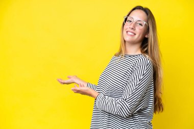 Sarı arka planda izole edilmiş genç beyaz kadın davet etmek için ellerini yana uzatıyor.