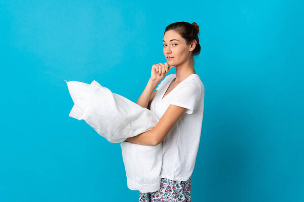 Молодая ирландская женщина изолирована на синем фоне в пижаме и держит подушку, думая: