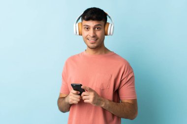 Genç Kolombiyalı bir adam mavi arka planda izole edilmiş. Cep telefonuyla müzik dinliyor ve ön tarafa bakıyor.