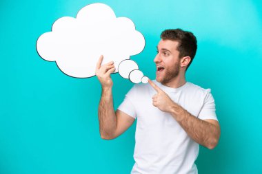 Kafkasyalı genç adam mavi arka planda izole edilmiş bir konuşma balonunu tutuyor ve ona doğrultuyor.