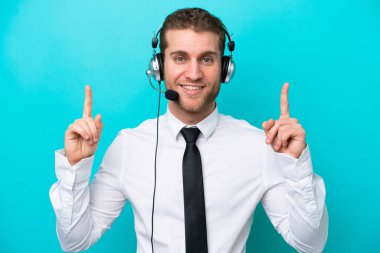 Tele pazarlamacı beyaz adam mavi arka planda izole edilmiş kulaklıkla çalışıyor. Harika bir fikre işaret ediyor.