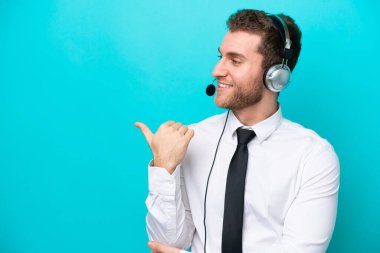 Tele pazarlamacı beyaz bir adam mavi arka planda izole edilmiş bir kulaklıkla çalışıyor. Yan tarafı göstererek bir ürün sunuyor.