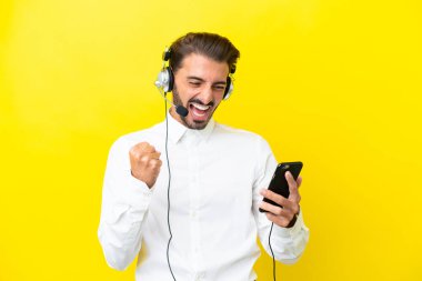 Telefon pazarlamacısı beyaz adam sarı arka planda izole edilmiş bir kulaklıkla çalışıyor ve telefon zafer pozisyonunda.