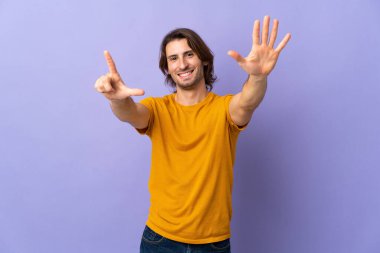 Mor arka planda izole edilmiş genç yakışıklı yediyi parmaklarıyla sayıyor