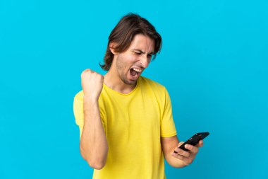 Mavi arka planda izole edilmiş genç yakışıklı cep telefonu kullanıyor ve zafer jesti yapıyor.