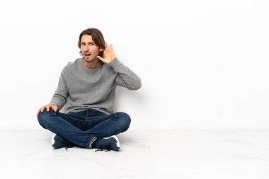 Genç yakışıklı adam izole edilmiş arka planda yerde oturuyor. Kulağına elini sürerek bir şeyler dinliyor.