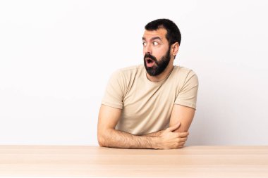 Masada sakallı beyaz bir adam yan tarafa bakarken sürpriz jest yapıyor..