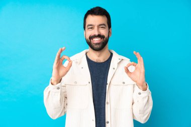 Mavi arka planda beyaz kadife ceketli genç ve yakışıklı bir adam parmaklarıyla iyi bir işaret gösteriyor.