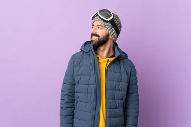 İzole edilmiş mor arka planda snowboard gözlüklü kayakçı yan bakıyor ve gülümsüyor.