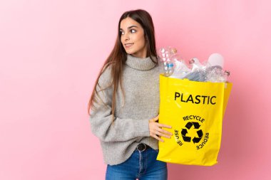 Pembe arka planda izole edilmiş ve gülümseyen plastik şişelerle dolu bir çanta taşıyan beyaz bir kadın.