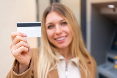 Genç, güzel, sarışın bir kadın dışarıda kredi kartı tutuyor.