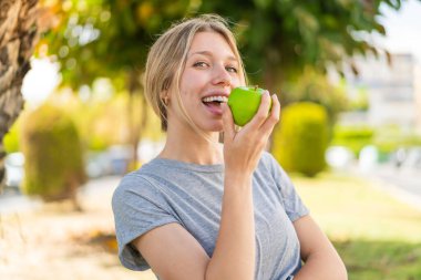 Açık havada elma yiyen genç sarışın kadın.