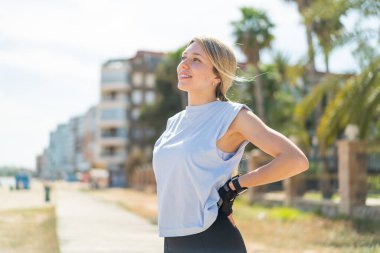 Açık havada genç sarışın bir kadın spor giyer ve neşeli bir ifade takınır.