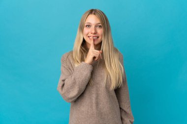 Mavi arka planda izole edilmiş genç sarışın bir kadın sessizlik işareti gösteriyor. Parmağını ağzına sokuyor.