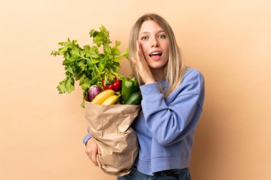 Bir çanta dolusu sebzeyi soyutlanmış arka planda tutan genç sarışın kadın bir şeyler fısıldıyor