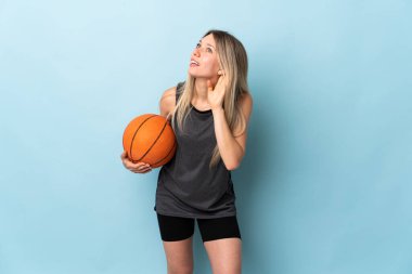 Basketbol oynayan genç sarışın kadın mavi arka planda izole edilmiş bir şekilde bir şeyler dinliyor.