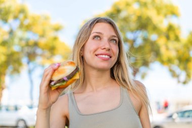 Sarışın bir kadın elinde hamburgerle yukarı bakıp gülümsüyor.