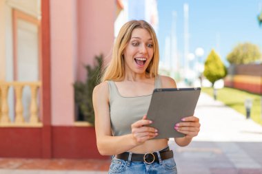 Açık havada genç sarışın bir kadın elinde tabletle şaşırmış bir ifadeyle.